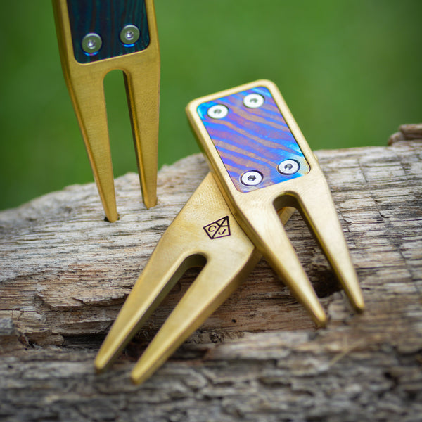 Three brass divot tools with blue, purple, and orange damascus inserts on a wooden surface with a green blurred background.